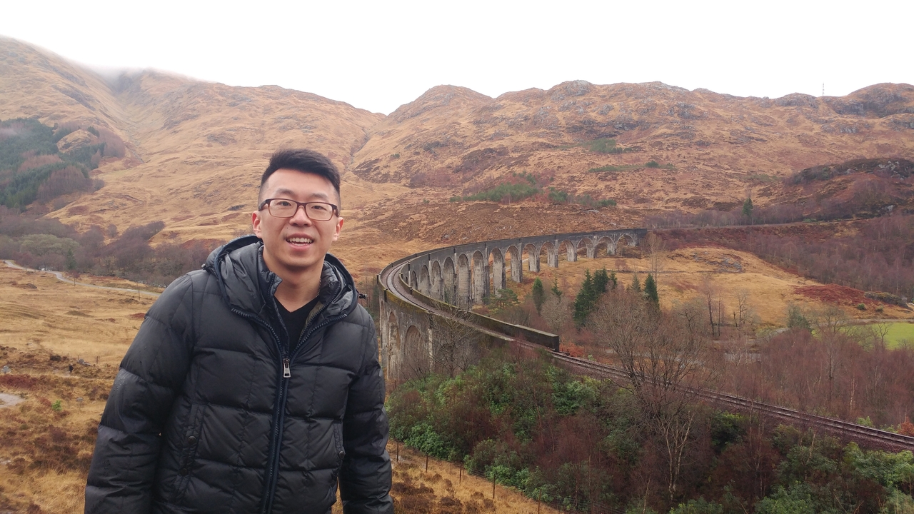 A person standing in front of the Glenfinnan Viaduct with scenic hills in the background.