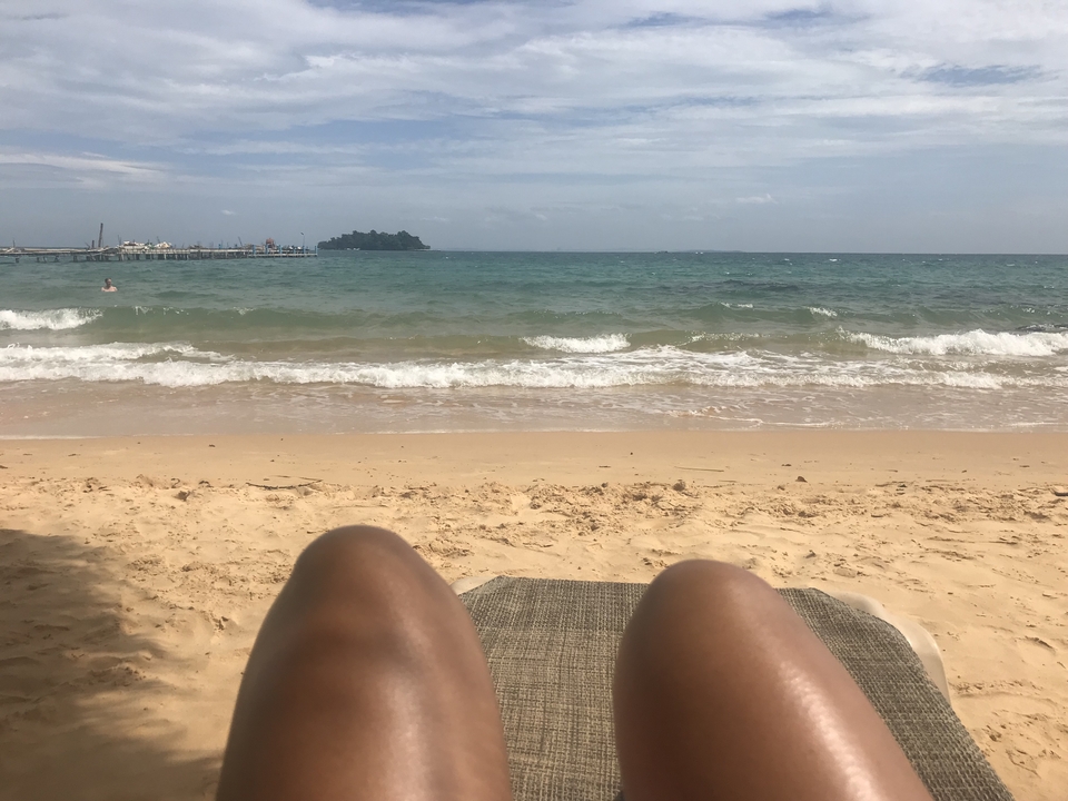 A beach with waves and an ocean view, with someone's legs in the foreground.
