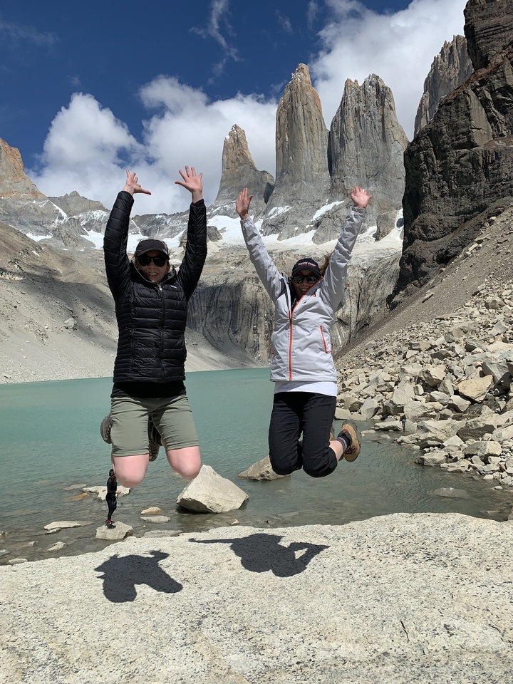 Two people jumping with a glacial lake and snowy mountain backdrop.