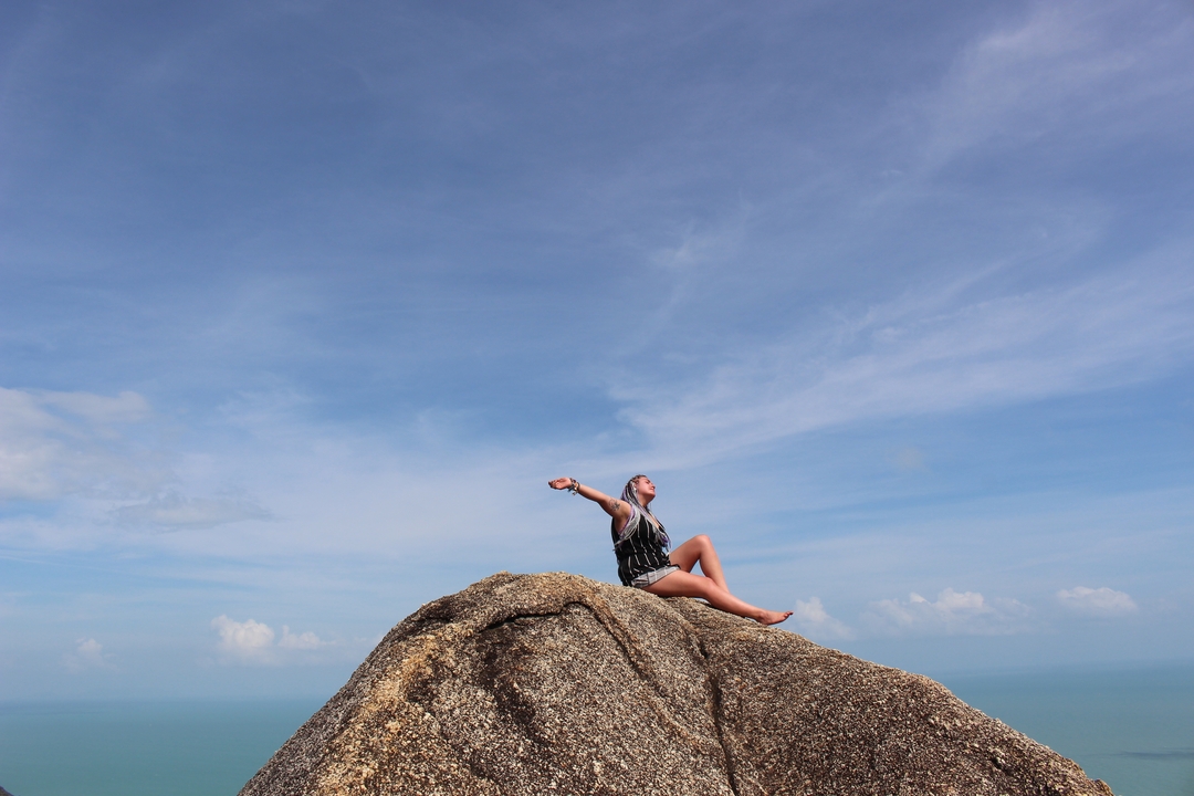 Person sitting on a rock with arms raised, blue sky.
