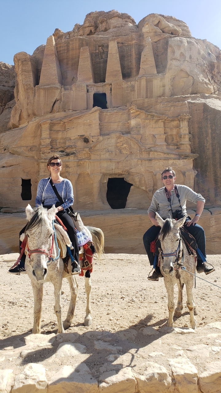 Two people riding camels with ancient rock-cut architecture in Petra.