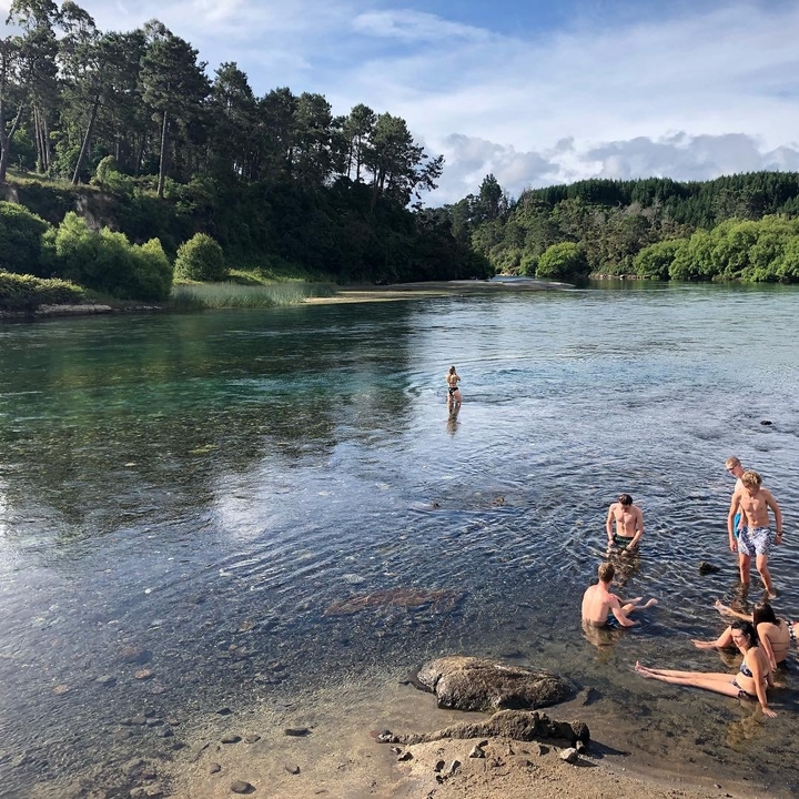 People swimming in a shallow clear river surrounded by green trees.