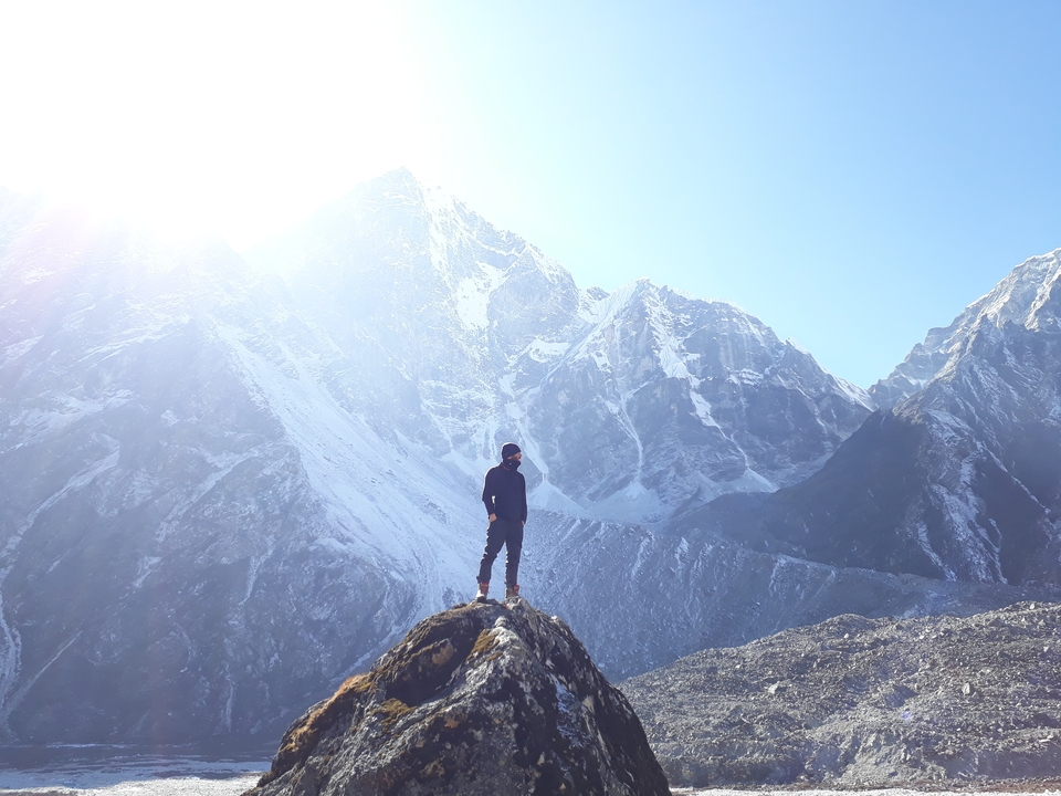 Personne debout sur un sommet rocheux avec des montagnes enneigées en arrière-plan.