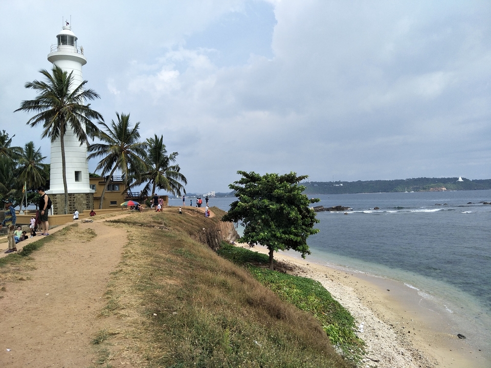 Lighthouse by a beach with people walking around.