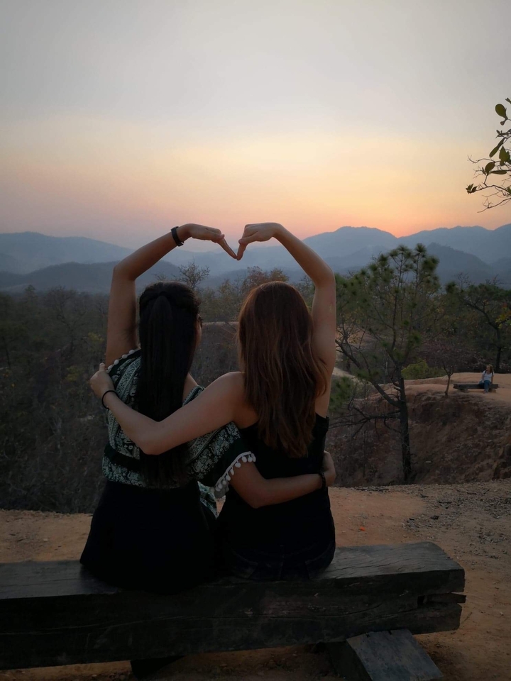Two people forming a heart shape with their hands overlooking a sunset.