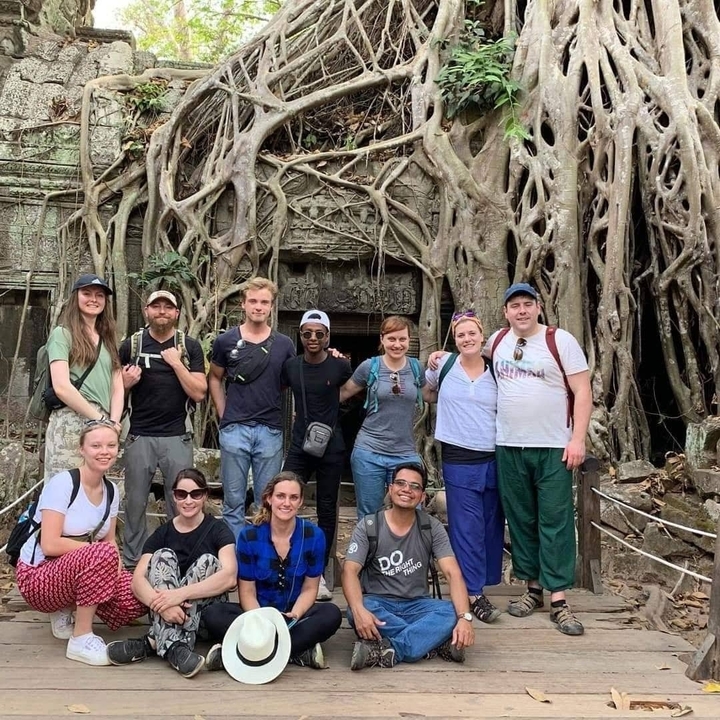 Group of tourists in front of tree roots covering temple ruins.