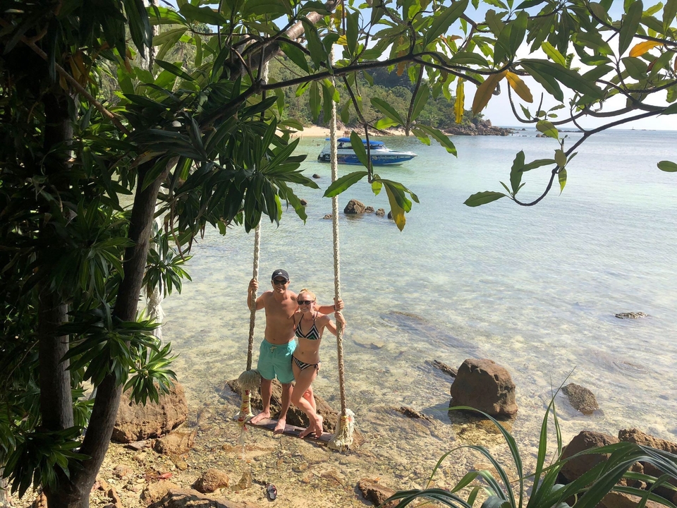 Couple on a swing by the beach with a boat in the background.