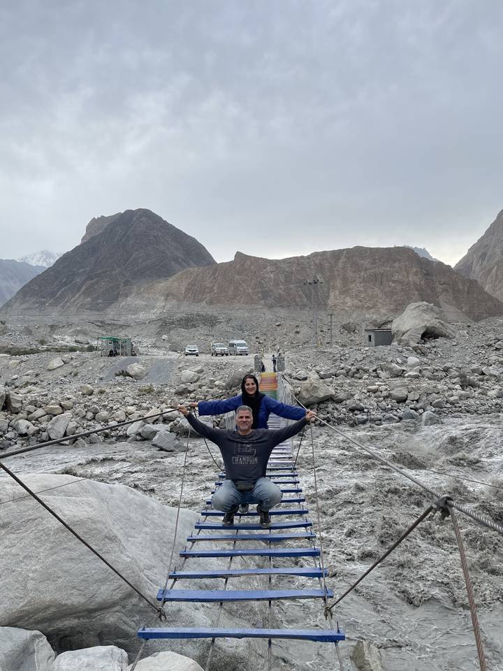Two people on a suspension bridge with rocky mountains behind.