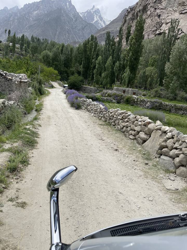 Rugged road with vehicle mirror, surrounded by trees and mountains.