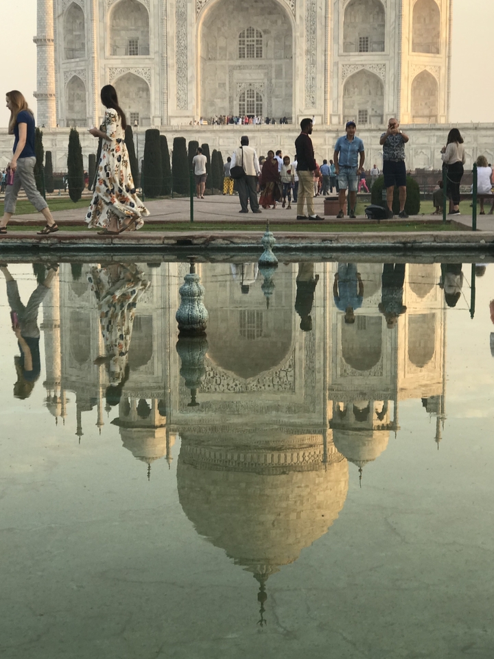 Reflet d'un mausolée blanc dans l'eau, avec des personnes qui marchent à proximité.