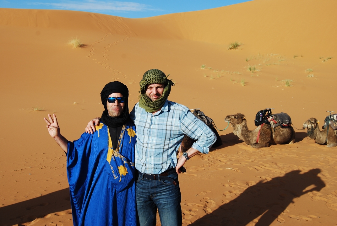 Two men posing with camels on sand dunes.