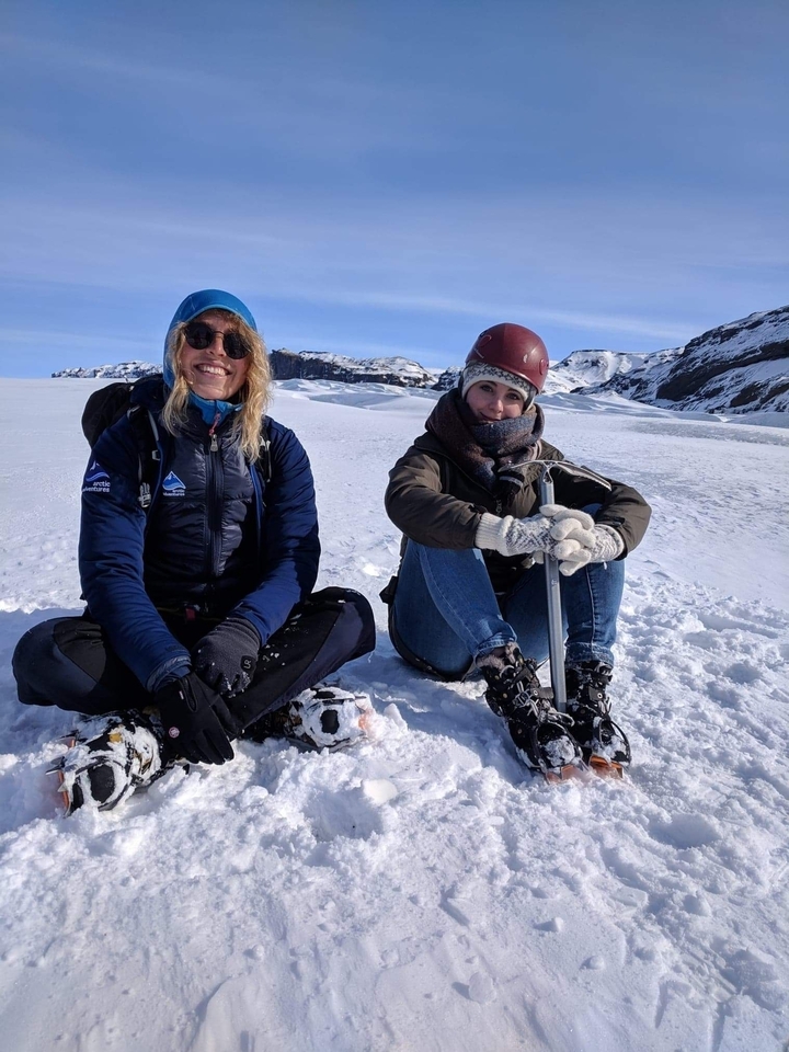 Two people sitting together on snow with mountains in the background.