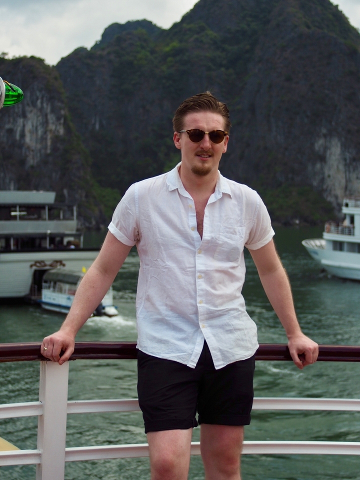 Man in a white shirt standing on a boat with cliffs behind him.