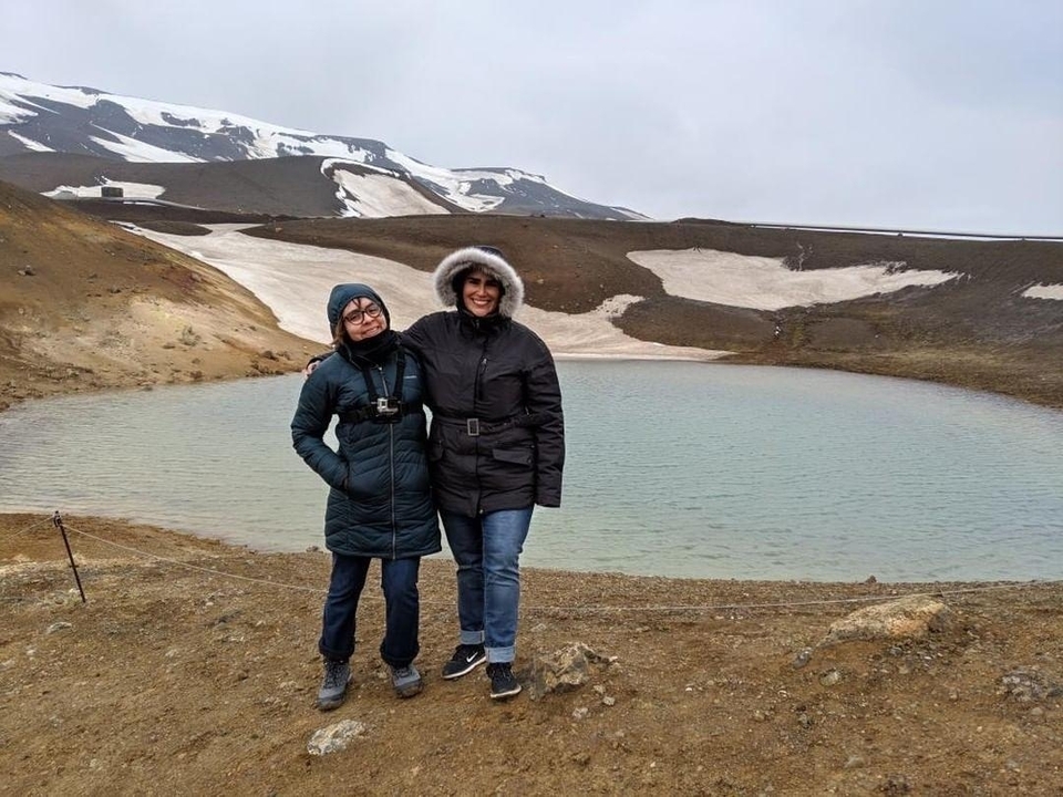 Two people standing by a scenic pond with snow patches and mountains in the background.