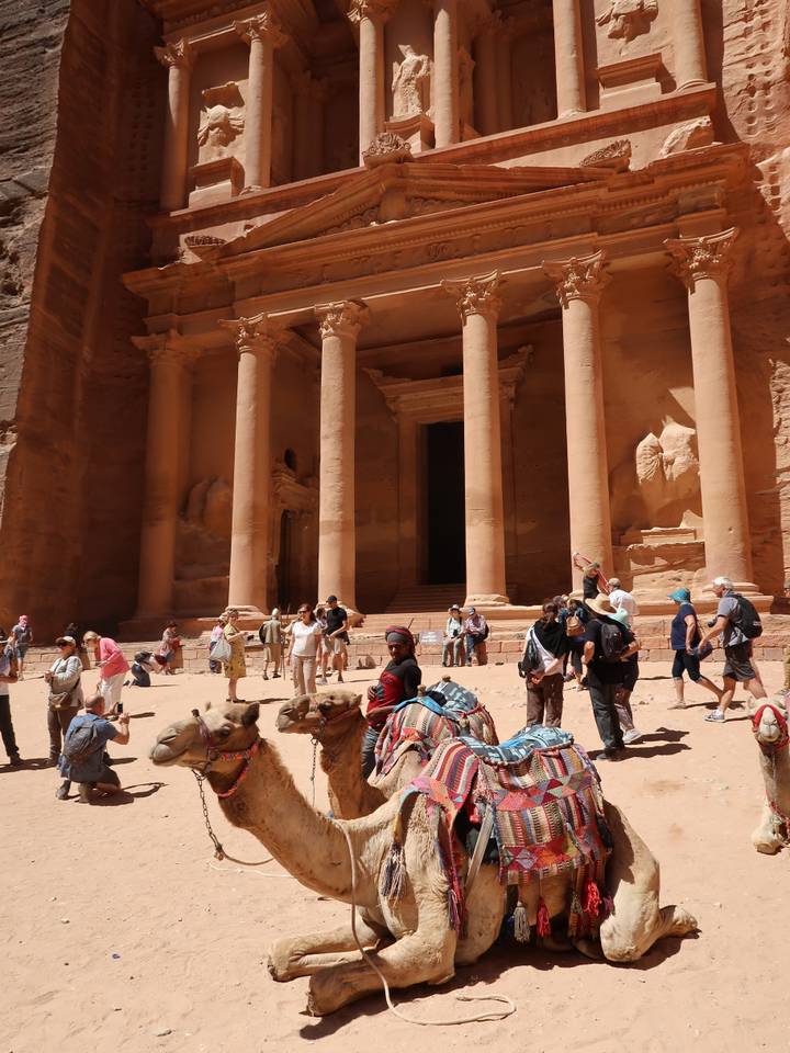 Visitors and camels in front of a grand, ancient rock-carved facade.