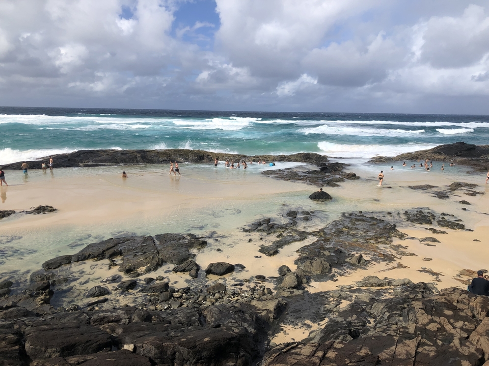 People enjoying natural tidal pools along a beach with crashing waves.