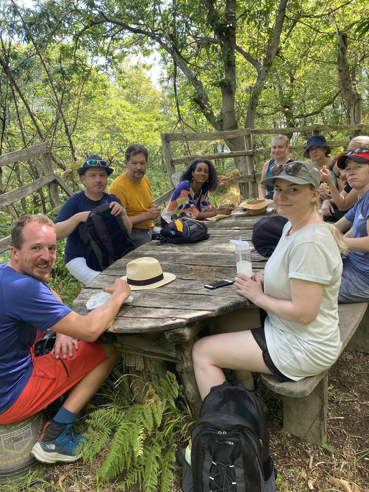 Group of people sitting at a picnic table in a wooded area.