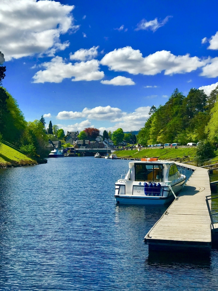 A scenic view of a canal lined with boats and a walkway, surrounded by lush greenery.