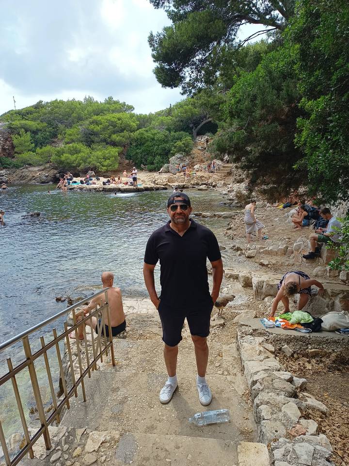 A man posing at a rocky beach with people in the background.