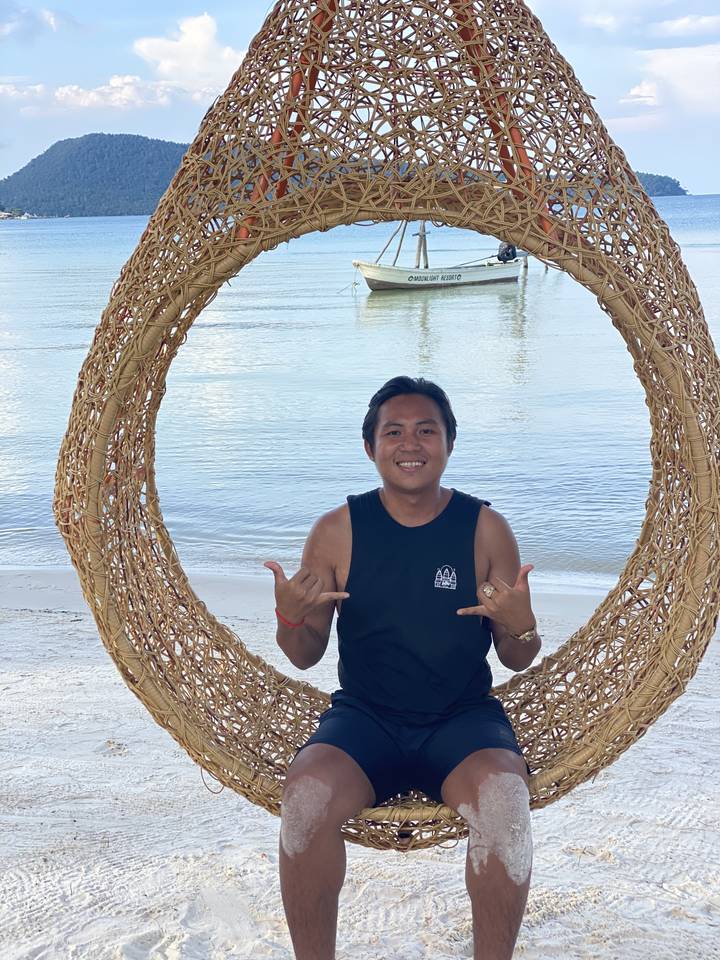 A person sitting on a wicker swing by the beach with a boat in the background.