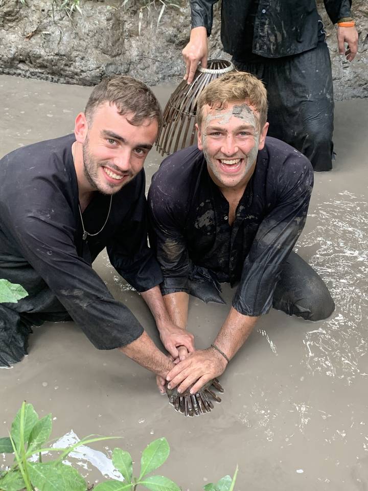 Two men smiling with muddy faces in water, posing with a crab trap.