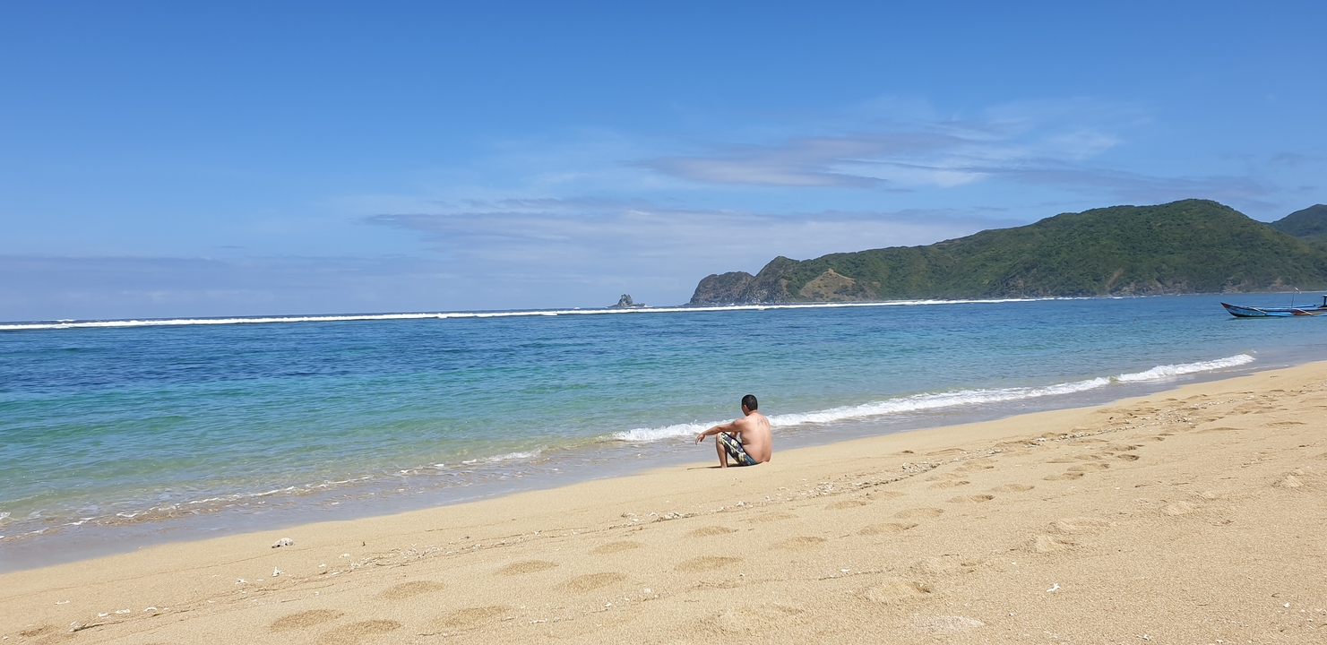 Une personne assise sur une plage de sable avec des eaux bleues claires et des collines lointaines.
