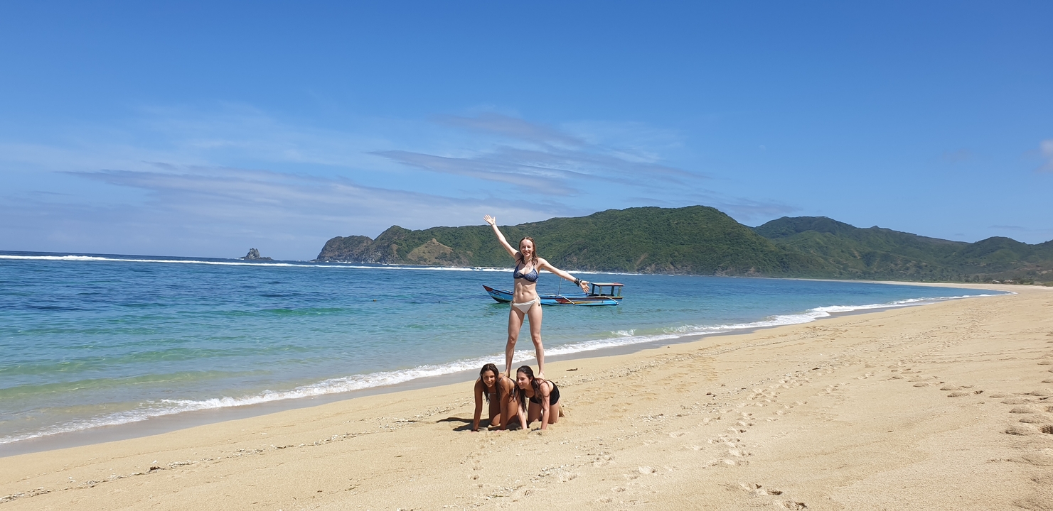 Trois personnes formant une pyramide humaine sur une plage de sable avec un bateau en arrière-plan.
