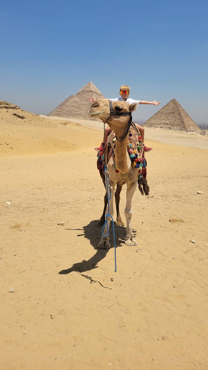 Person riding a camel in the desert with pyramids.