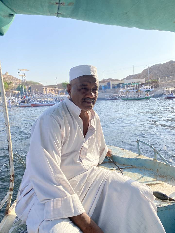Man sitting in a boat on a river with other boats on the opposite bank.