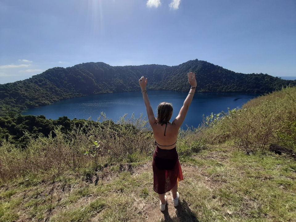 Person with arms raised viewing a crater lake and forested hills.