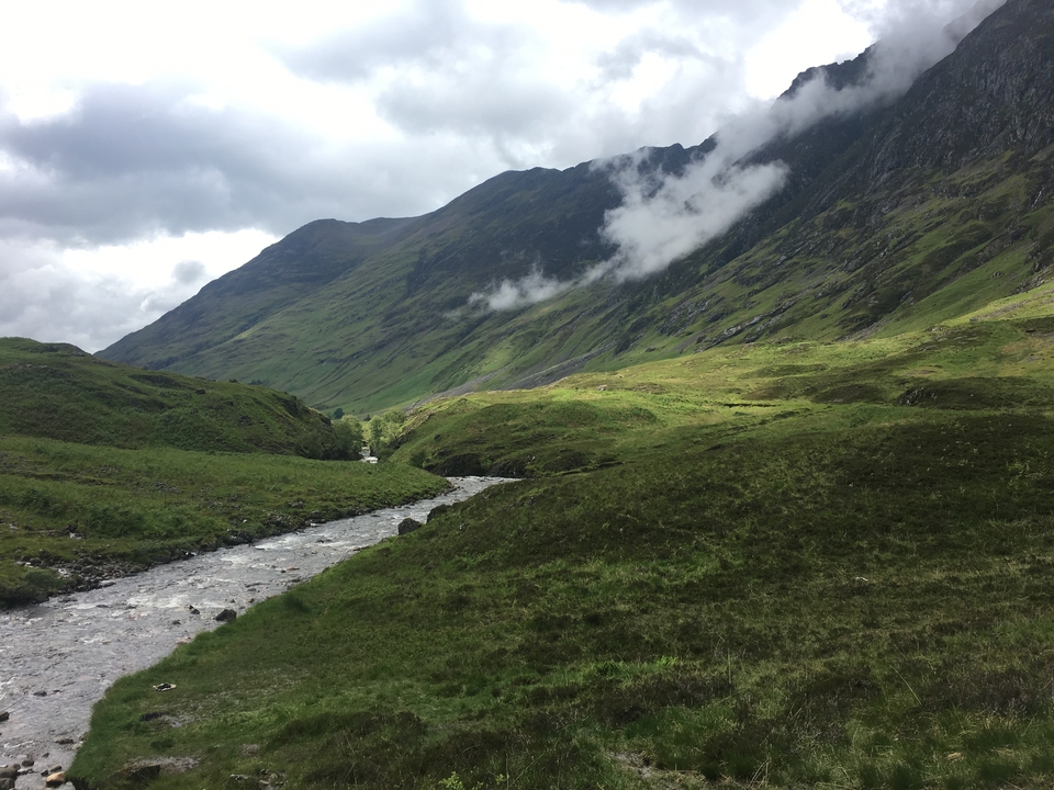 Rolling green hills and a stream under a cloudy sky.