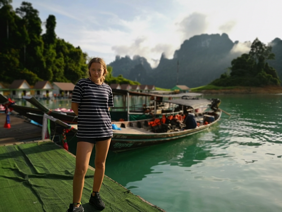 Person standing on a dock with boats and mountains in the background.