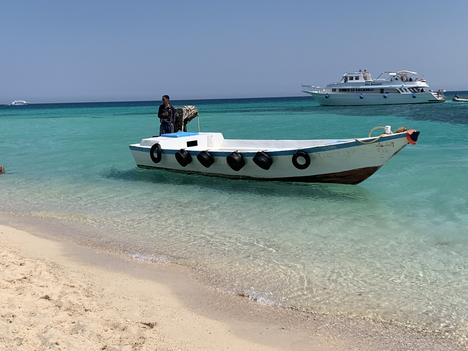 Man standing on a small boat near a sandy shore with clear water.
