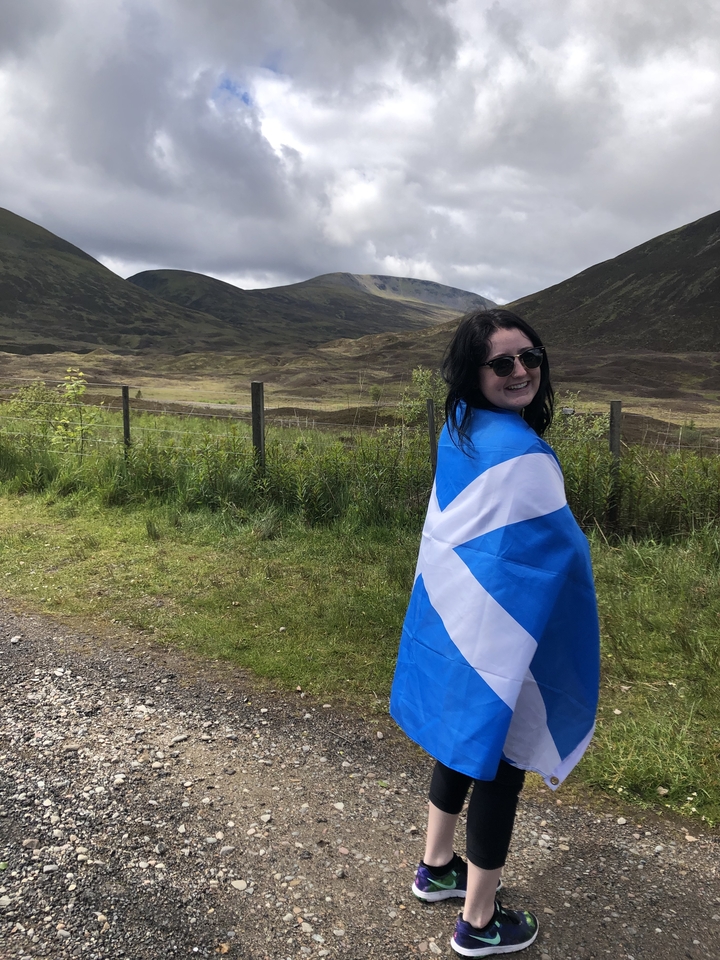 A person wrapped in a flag standing by a fence in a vast landscape.
