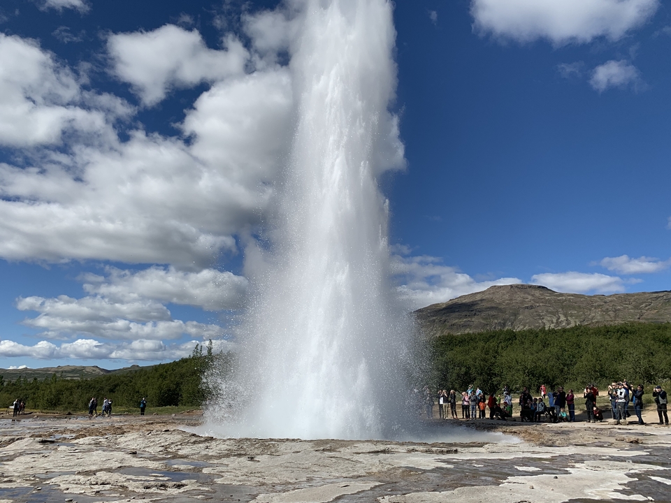Geyser eruption with a crowd of onlookers and mountains in the background.