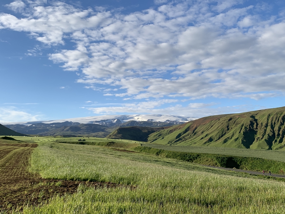 Wide expanse of green fields with distant snow-covered mountains under a cloudy sky.
