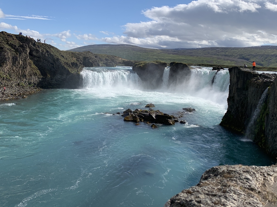 Waterfall with people standing at its edge and rocks below.