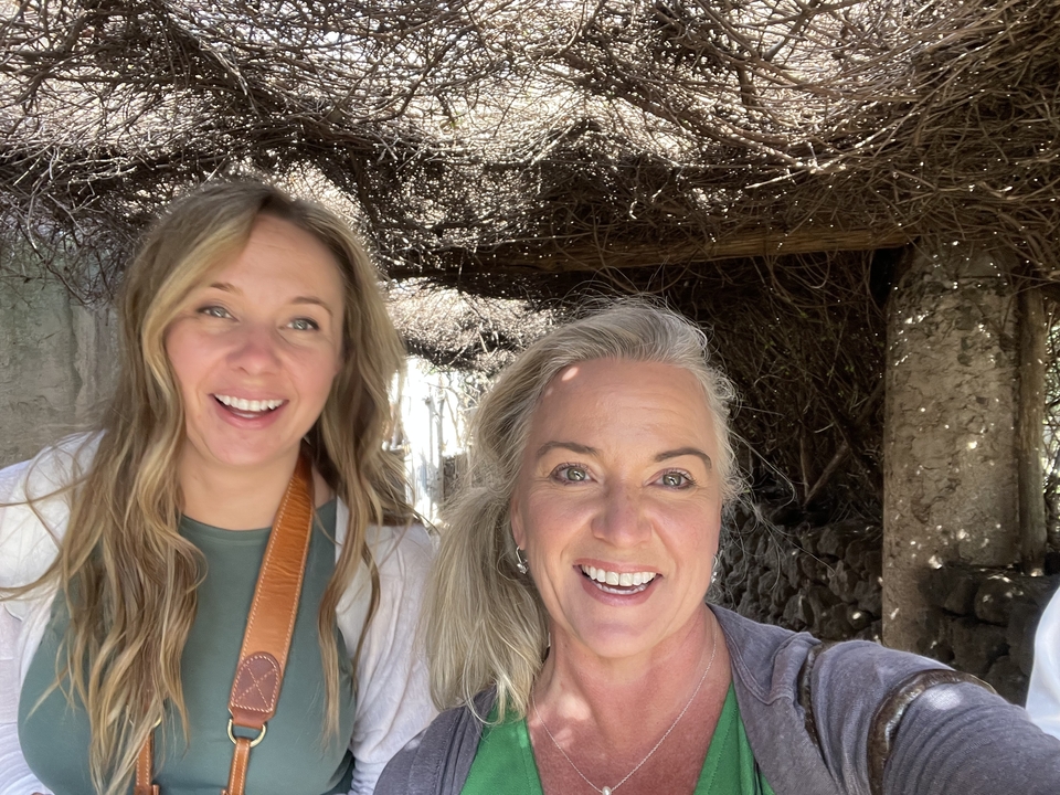 Two smiling women under a vine-covered canopy.