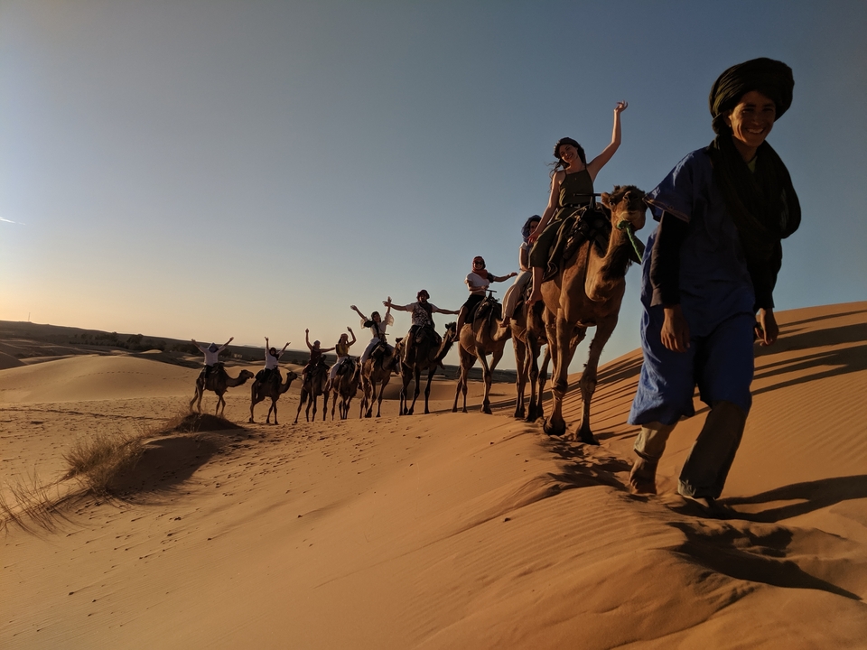 People riding camels in a desert landscape during sunset.