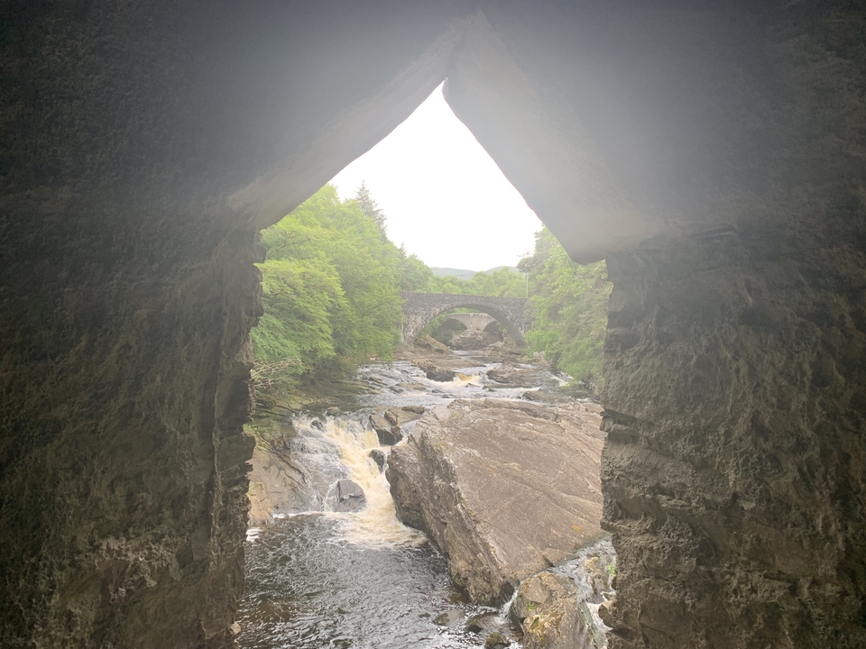 Stone arch bridge over a flowing river with greenery.