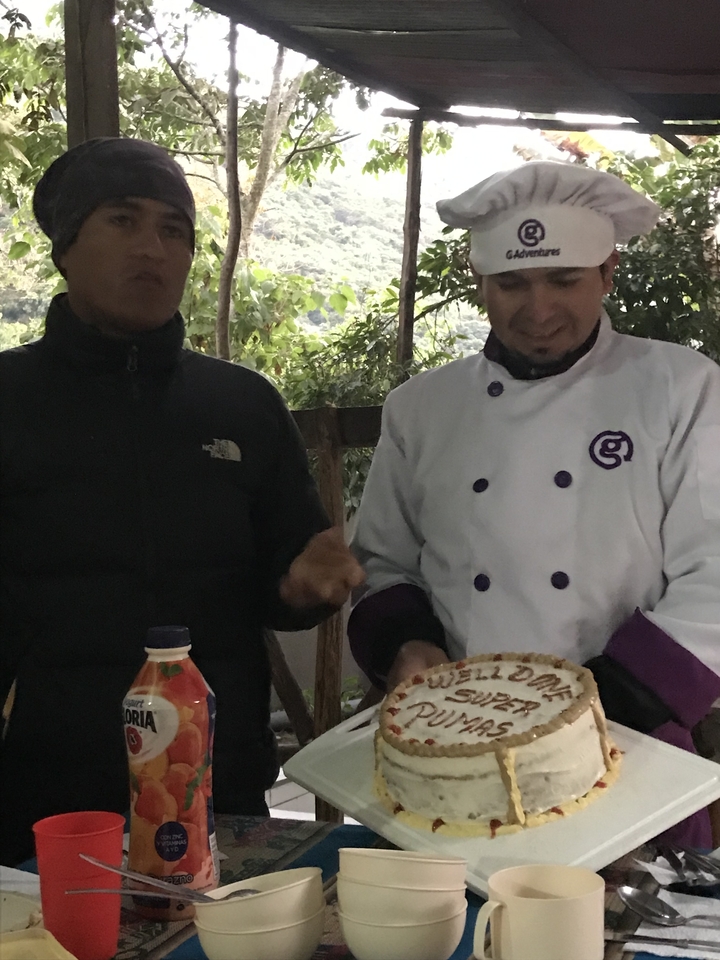 Dos hombres presentando un plato en un entorno al aire libre boscoso.