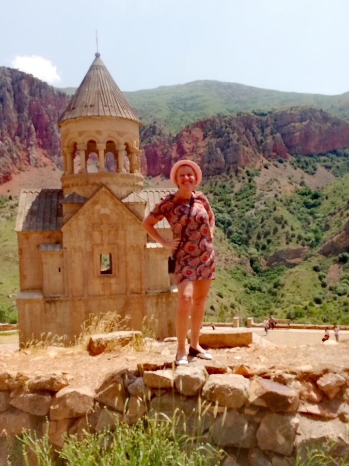 Smiling woman in patterned dress and hat posing beside a small stone church set against dramatic red cliffs and green valley.