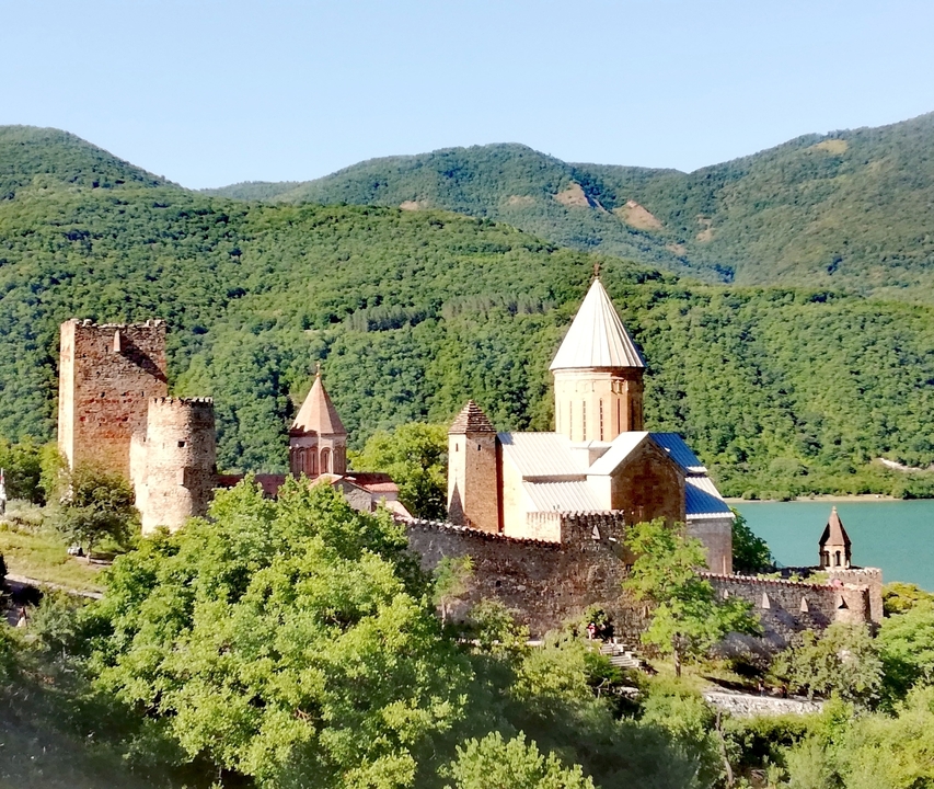Historic Ananuri fortress complex with stone towers and conical-roofed church beside emerald reservoir and forested mountains.