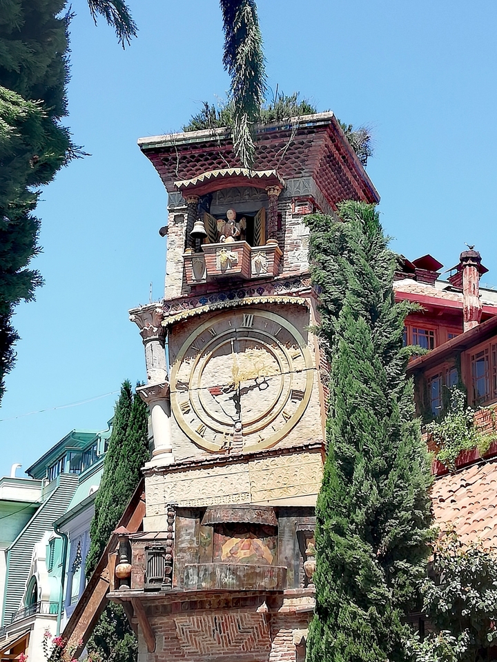 Whimsical leaning clock tower of the Rezo Gabriadze Theatre framed by tall cypress trees under a bright blue sky.