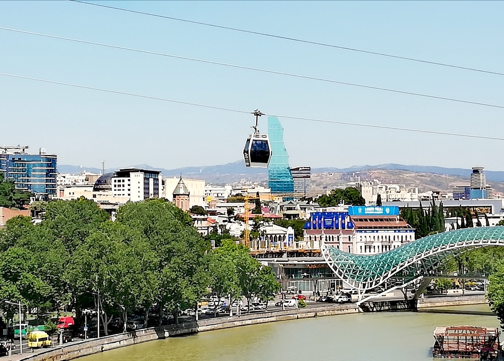 Cityscape of Tbilisi with cable car cabin gliding above trees, modern buildings and the glass Bridge of Peace in the foreground on a clear day.