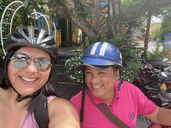 Two people wearing helmets, smiling with a street in the background.