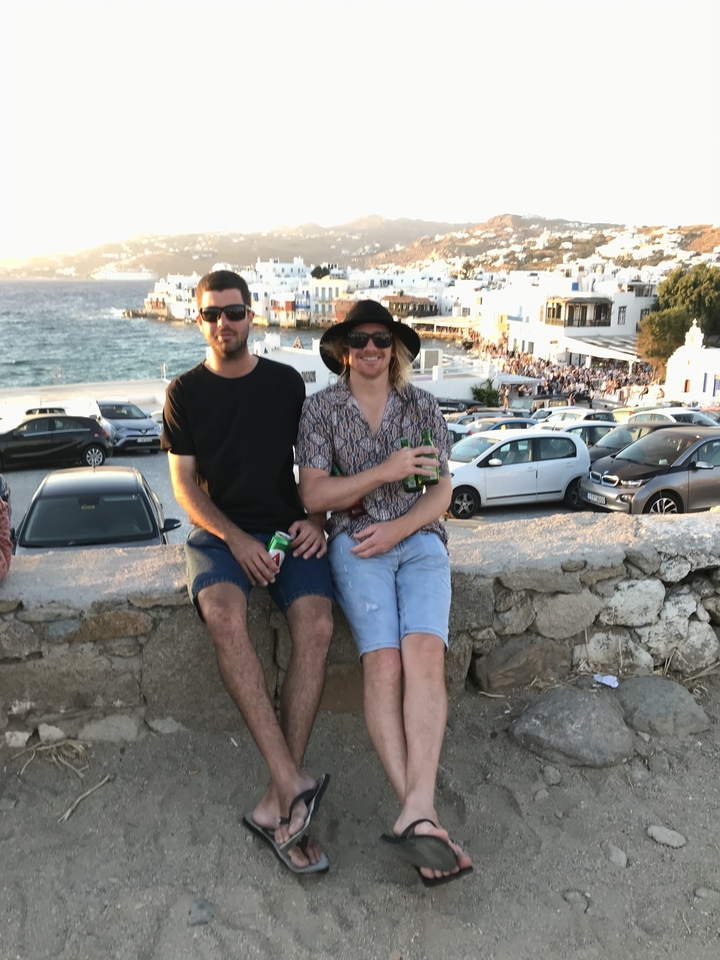 Deux hommes assis sur un mur surplombant une scène côtière avec des voitures garées.