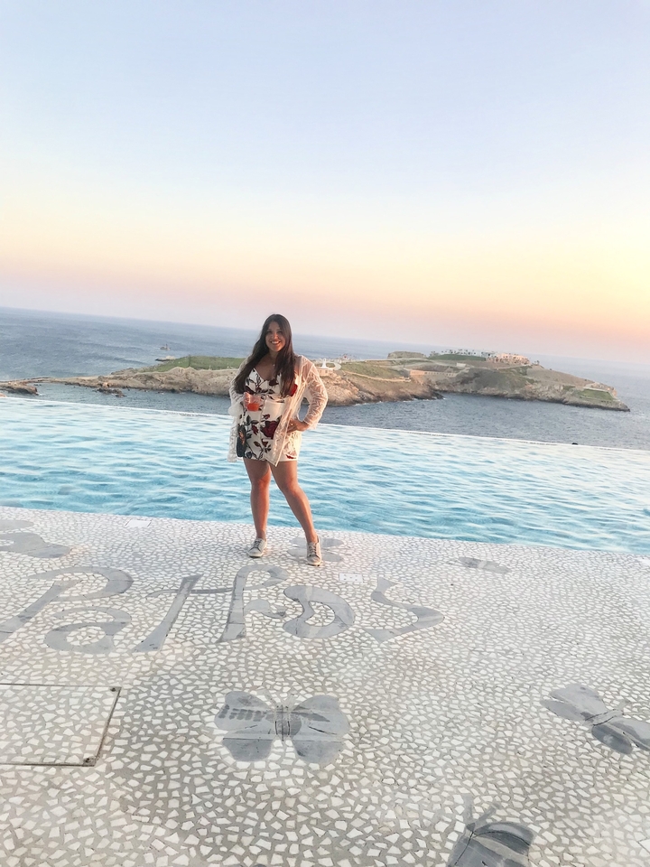 Une femme posant au bord d'une piscine avec une vue panoramique sur l'océan au coucher du soleil.