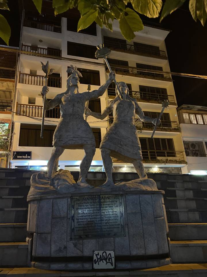 Statue de pierre de deux personnages avec un arrière-plan de bâtiments modernes.