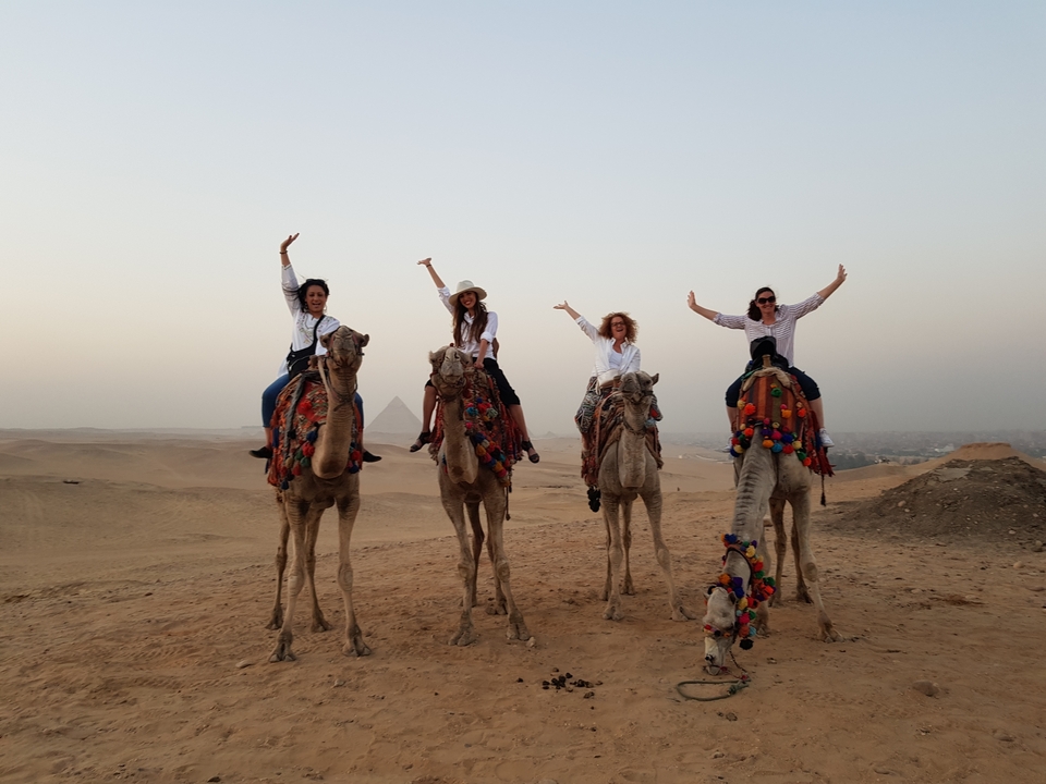 Group of women riding camels in a desert with pyramids in the background.